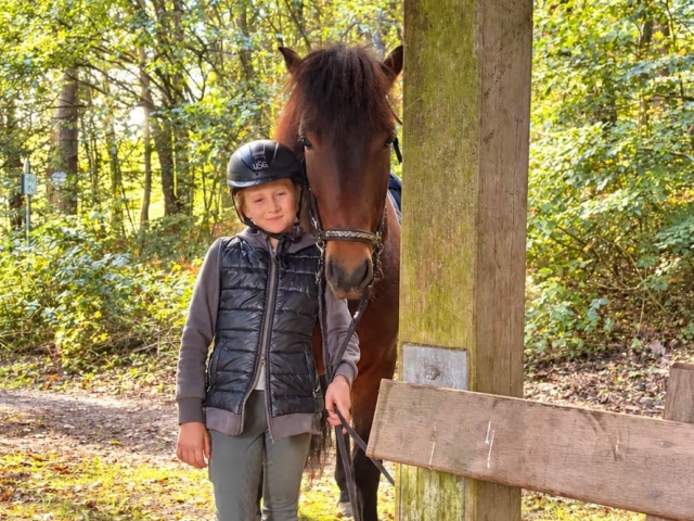 whatsapp image 2026 10 04 at 20.18.15 Kind mit Reithelm steht neben braunem Pferd im Wald